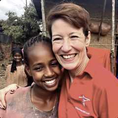 A smiling woman in a red shirt hugs a smiling young girl outdoors, with another girl standing in the background near a rustic building.