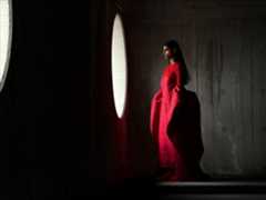 A woman wearing a long red dress stands in an empty room being photographed. The Photo app toggles through the focal lengths to demonstrate the capabilities of the pro camera system, showcasing: 100 mm