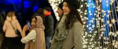 People in coats smile broadly under a tunnel of tiny lights outdoors at night.