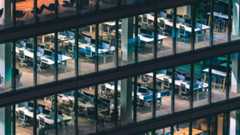 An exterior view of an office building with large glass windows and empty tables and desks inside