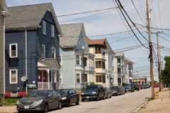 A photo of a street with triple decker houses on the left side and with cars parked in front of the houses.