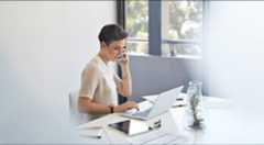 オフィスの机で、スマートフォンで話しながらノートパソコンを操作するショートヘアの女性