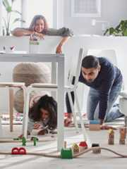 ダイニングテーブルの下で大人が膝をついて子どもと遊んでいる。その後ろに大人がもう1人いる。