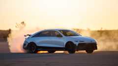 A white Ioniq 6 N drifting on asphalt in the desert creating a cloud of smoke in front of a sunset with Joshua trees in the background