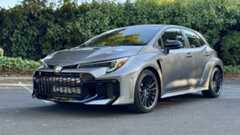 A front three-quarters shot of a silver GR Corolla in a parking lot in front of hedges