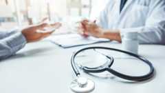 Arztbesuch Zwei Personen sitzen am Schreibstisch. Im Vordergrund liegt ein Stetoskop.