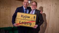 Die Oberbürgermeister von Kyjiw und Leipzig im Februar 2025 Zwei Herren umarmen sich mit Ortsausgangsschild von Leipzig vor einer hölzernen Wand.