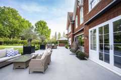Florence Grey outdoor tiles UK stretch across width of house, with 2 sets of rattan furniture, olive tree planters and topiary.***Elitescapes, www.elitescapes.co.uk