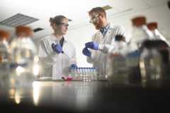 A student and professor work on a science research project together. They are wearing white lab coats and goggles while they hold lab equipment.