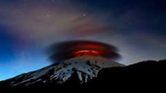 A double cloud formation is lit up at night by the lava emitted from the top of a volcano.