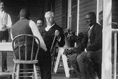 Men in dressed in suits and waistcoats sit on the porch of large building.