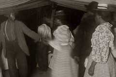 A man and woman hold hands as they dance. Others look on and some look towards the camera.