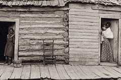 A woman holds a baby and a young girl stares straight on in the doorways of a log cabin.