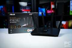 The black ASUS RT-BE58U (BE3600 router sitting on a white table with a background of blurry PC component boxes on a bookshelf. It sits in front of its box with four antenna sticking up from it.