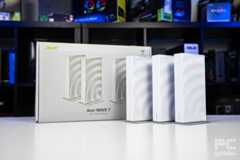 The three Acer Wave 7 routers standing next the their box sitting on a white table with a background of blurred PC component boxes on a bookshelf.