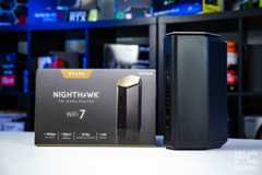 The Netgear Nighthawk RS600 with its box standing on a white table, with a background of a bookshelf filled with blurred PC component boxes.