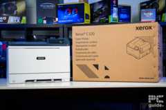 The Xerox C320 front on, on the left side, and its box on the right of it. Sitting on a white table, with a background of a bookshelf filled with PC component boxes.