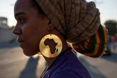 This profile shows a young protestor wearing a large, round, gold earring with a cutout of the African continent and two multicolored headwraps.