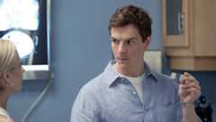 Dexter (Patrick Gibson), wearing a blue button-down shirt and a serious expression, holds up a microscope slide as he speaks to a blonde woman in a lab on Dexter